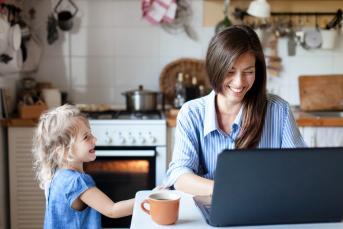 Junge Frau sitzt an ihrem Laptop in der Küche. Neben ihr steht ihre kleine Tochter.